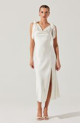 A woman is wearing a long, white, elegant dress with shoulder straps tied into bows. The dress has a cowl neckline and a side slit that reveals her leg. She is also wearing open-toed, high-heeled sandals and gold hoop earrings.